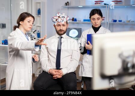 I ricercatori neurologici spiegano il risultato del trattamento puntando sul monitor mentre lo scienziato medico agguutaking le note, la scansione delle cuffie si prepara per la scansione del cervello che analizza l'attività elettrica. Foto Stock