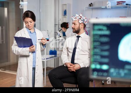 Medico che parla di sintomi di malattia durante le indagini utilizzando cuffie high tech per la scansione delle onde cerebrali. Ricercatore che analizza lo stato di salute del paziente umano, funzioni cerebrali, sistema nervoso. Foto Stock