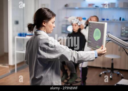 Neurologo medico analizzando la scansione cerebrale sul display del monitor nel moderno laboratorio di ricerca, sviluppando una cura per la diagnosi del sistema nervoso. Paziente donna che indossa cuffie con sensori Foto Stock