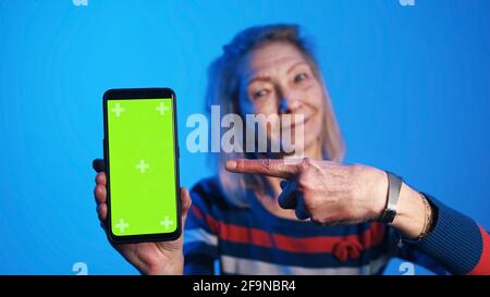 Donna anziana che tiene uno smartphone con schermo verde verticale e punta il dito verso di esso con un sorriso. Foto di alta qualità Foto Stock