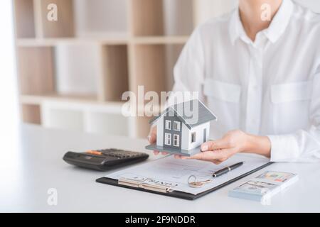 Affari. L'uomo in una camicia bianca porta un modello di casa. Su un tavolo ha carte commerciali, calcolatrice, penna, chiave di casa e banconote. Foto Stock
