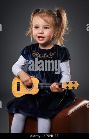 Adorabile ragazza bionda che suona la chitarra, studio girato su sfondo grigio Foto Stock