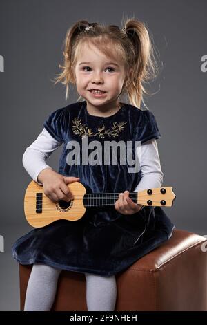 Adorabile ragazza bionda che suona la chitarra, studio girato su sfondo grigio Foto Stock