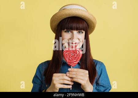 Primo piano di un'affascinante giovane donna dai capelli rossi, con cappello di paglia e camicia in jeans, che tiene un lollipop rosso dolce a forma di cuore, sorridendo alla telecamera su uno sfondo giallo. Foto Stock