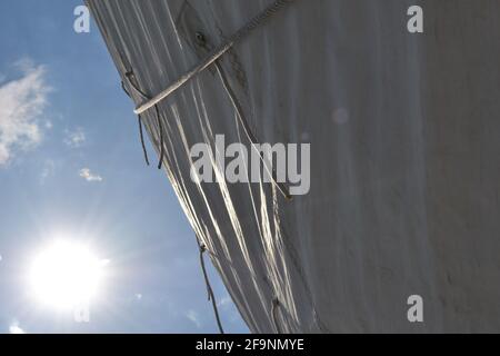 Il sole che brilla sulla vela bianca di una barca; raggi di sole luminosi in un cielo blu, sopra la vela e i punti della scogliera della corda Foto Stock