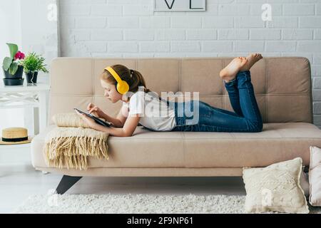 Bambina dai capelli rossi che si stette sul divano con un tablet elettronico ascolta la musica in cuffia. Foto Stock