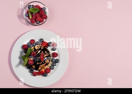 Vista dall'alto sui pancake americani fatti in casa con mirtilli freschi, lamponi, fragole e sciroppo di cioccolato. Colazione sana al mattino. Rosa b Foto Stock
