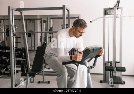 Un uomo stanco in una T-shirt bianca esegue esercizi su una cyclette. Allenamento cardio. Foto Stock