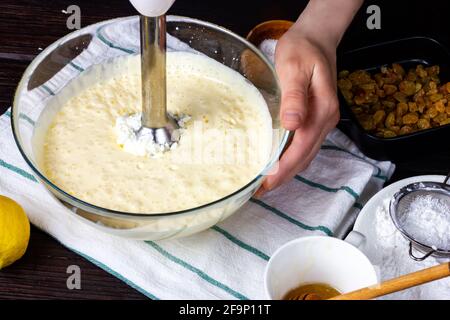 Il processo di impastare una pastella per una casseruola cagliata. Mani femminili che tengono una ciotola di vetro della pastella. Il frullatore montare il formaggio da cottage, il latte e. Foto Stock