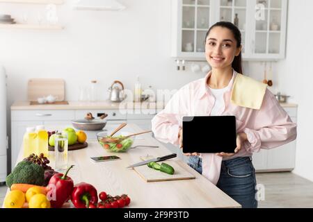 Donna che mostra tablet con schermo vuoto per mockup in cucina Foto Stock