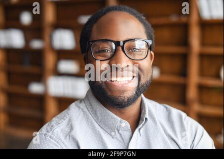 Primo piano ritratto di un felice afro-americano giovane con amichevole ampio sorriso toothy, un ragazzo bearded razza mista indossando eyeglasse elegante e camicia guarda in macchina fotografica, foto del profilo del dipendente Foto Stock