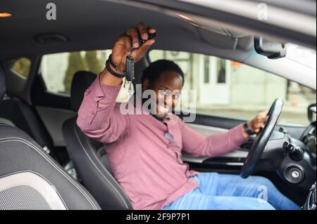 Fuoco selettivo alle chiavi dell'automobile in mano dell'uomo afro-americano felice. Allegro ragazzo nero che acquista una macchina, ha ottenuto una patente di guida, si siede e tiene il volante e guarda la macchina fotografica, raggiungendo l'obiettivo Foto Stock