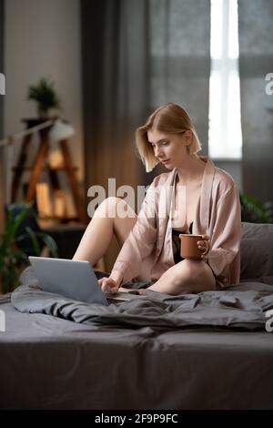 Giovane donna seduta sul letto con una tazza di caffè e digitando sul computer portatile al mattino Foto Stock