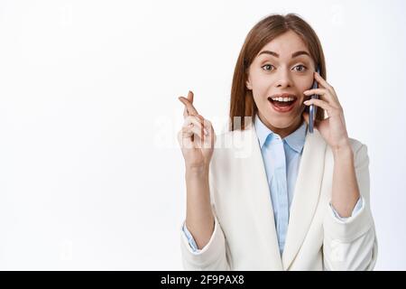 Sorridente donna d'affari in tuta parla al telefono con le dita incrociate, attende buone notizie di omen, parlando su smartphone con il volto allegro, in piedi Foto Stock