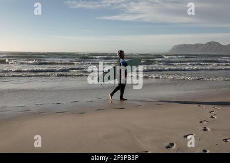 Donna afro-americana anziana che porta surf board mentre cammina sulla spiaggia. Concetto di vacanza di viaggio di riposo stile di vita Foto Stock