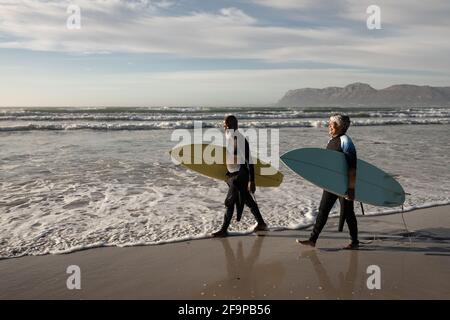 Coppia africana americana anziana che trasporta le tavole da surf che cammina sulla spiaggia. Concetto di lifestyle di pensione di vacanza di corsa Foto Stock