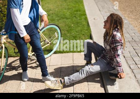 Due amici maschi di razza felici seduti su skateboard e in bicicletta per strada e parlare. Vita urbana verde, fuori e circa in città. Foto Stock
