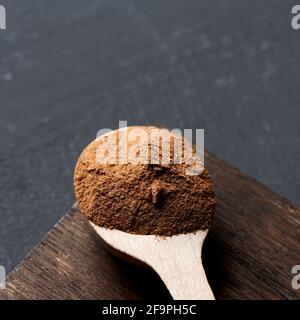 primo piano di un cucchiaio di legno pieno di polvere di camu-camu su un vassoio di legno scuro, posto su una superficie di grigio scuro, in un formato quadrato Foto Stock