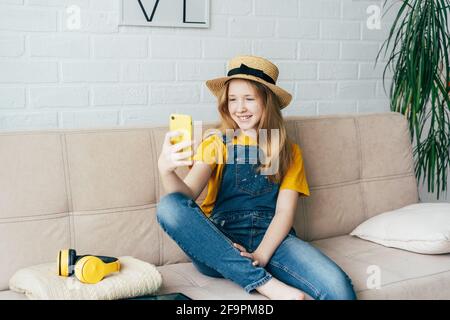 Allegra ragazza rossa in un cappello di paglia e tute in denim che posano e prendono un selfie su uno smartphone. Foto Stock