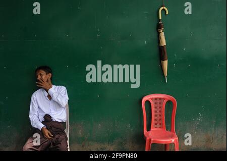 22.08.2013, Yangon, , Myanmar - UN uomo si siede accanto ad una sedia di plastica rossa e ad un ombrello appeso su un muro verde nell'ex capitale. 0SL130822D005CAR Foto Stock