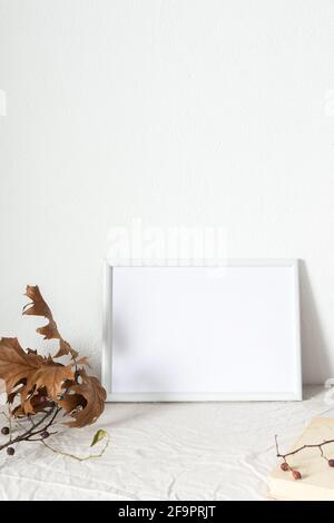 Casa decoratore moccap, quadro vuoto cornice vicino bianco dipinto muro di cemento, rami con foglie secche e libri antichi Foto Stock