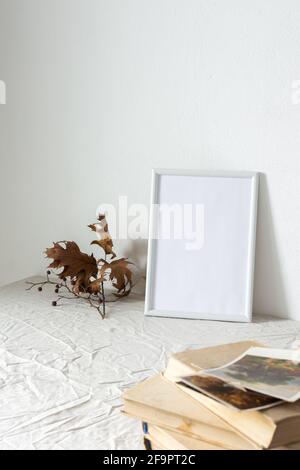 Casa decoratore moccap, quadro vuoto cornice vicino bianco dipinto muro di cemento, rami con foglie asciutte e vecchi libri con cartoline Foto Stock