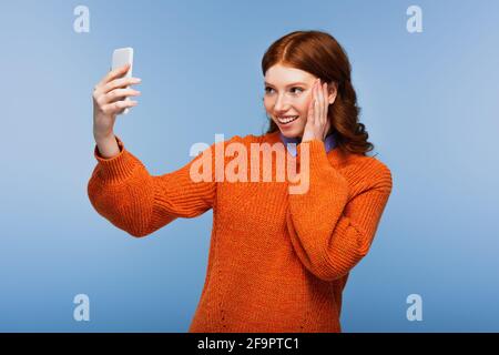 giovane donna sorridente in maglia arancione prendendo selfie isolato su blu Foto Stock