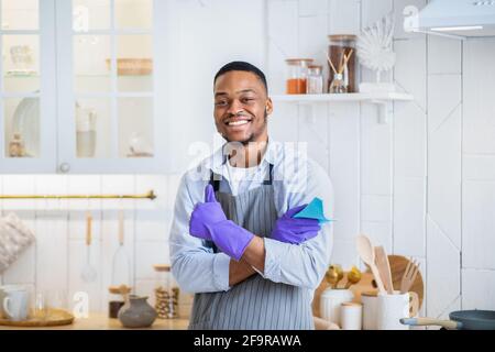 Bel giovane uomo nero in guanti di gomma sorridente alla macchina fotografica e mostrare il pollice in su gesto in cucina Foto Stock