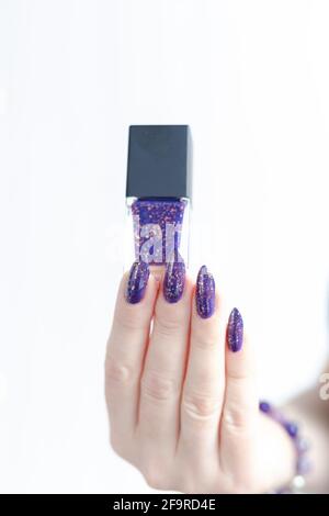 Mano femminile con unghie lunghe e manicure lilla viola tiene una bottiglia di smalto per unghie Foto Stock