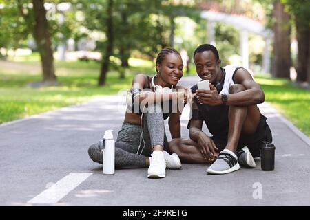 Coppie africane che usano l'app Fitness sullo smartphone insieme, riposandosi dopo l'allenamento all'aperto Foto Stock
