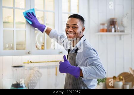 Ritratto di giovane ragazzo nero in guanti di gomma che si stigura cucina armadio e mostrare il movimento del pollice verso l'alto a casa Foto Stock