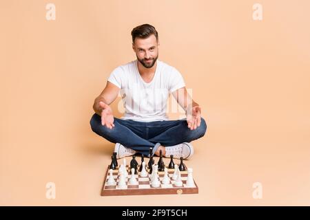 Ritratto di simpatico ragazzo acclamato seduto a terra a giocare a scacchi invitandoli ad unirsi isolato su sfondo di colore pastello beige Foto Stock