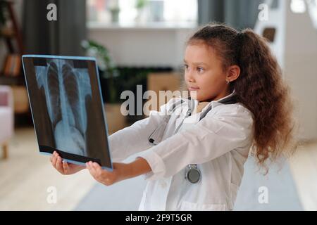 Bambina in camice bianco che tiene l'immagine a raggi X e che esamina lei gioca in ospedale a casa Foto Stock