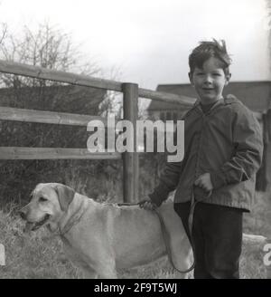 1967, storico, su un sentiero di campagna, un ragazzo fuori che prende il suo cane da compagnia, un Labrador ritorante per una passeggiata. I Labrador sono un animale domestico popolare, essendo un compagno amichevole, leale, paziente e non sono aggressivi, grandi come un animale domestico di famiglia. Ci sono molti benefici per i bambini nella cura per un cane, compreso l'aiuto sviluppare la loro autostima, la responsabilità imparante e l'empatia. Ci sono anche gli aspetti di salute di camminare un cane all'esterno e di costruire un sistema immunitario più forte. Foto Stock