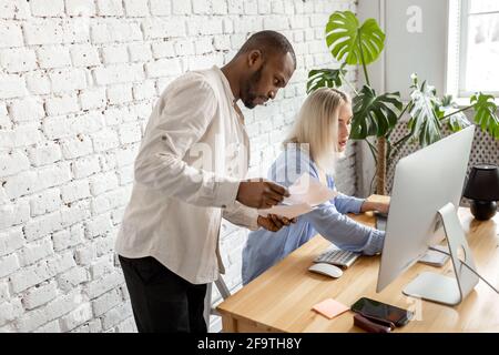 Colleghi di gara misti utilizzando il computer, facendo alcuni documenti, l'apprendimento di grafici finanziari. Giovani colleghi di stile che discutono nuove idee per il progetto Foto Stock