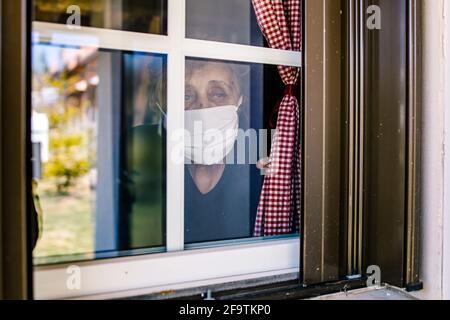 Donna anziana, nonna che indossa una maschera che guarda attraverso la finestra. Vista dall'esterno Foto Stock