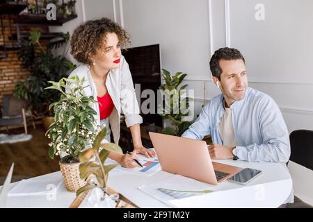 Partner aziendali concentrati che pensano alle idee di progetto, lavorando insieme in un ufficio moderno. Colleghi seri e alla moda che usano il laptop, imparando Foto Stock