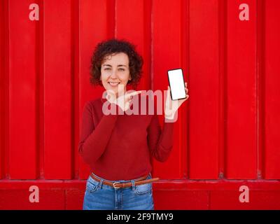 Una giovane donna caucasica con capelli ricci mostra il telefono alla macchina fotografica mentre sorride, indossando un maglione rosso e jeans e sullo sfondo c'è un Foto Stock