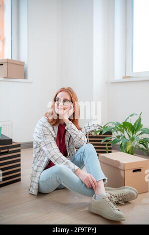 In un nuovo appartamento a luce bianca, una ragazza si siede sul pavimento e si pone sullo sfondo di scatole di cartone e un fiore in vaso. Foto Stock