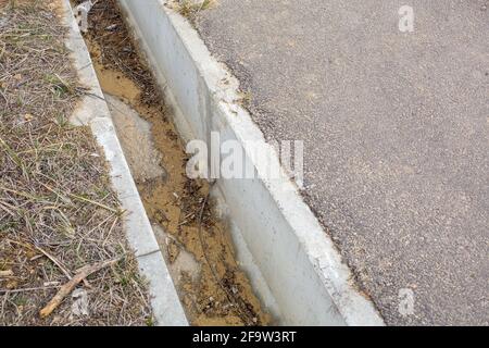 gocciolatoio di drenaggio in calcestruzzo sul lato di una strada asfaltata contaminata da detriti lavati dalle acque piovane da miglioramenti tecnici. Foto Stock