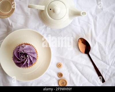 Cupcake con glassa viola e una teiera bianca con rosa cucchiaio d'oro su uno sfondo piuttosto bianco testurizzato Foto Stock