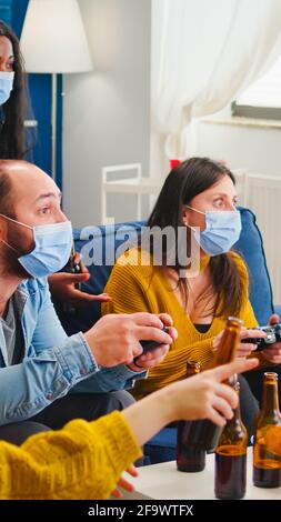 Amici tristi perché perdere di giocare ai videogiochi con controller wireless indossando la maschera facciale per evitare di ammalarsi con corona in tempo di pandemia sociale. Gruppo di persone che si divertono insieme. Foto Stock