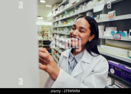 Allegro farmacista femminile che indossa camice da laboratorio alla ricerca di medicina in scaffale in farmacia Foto Stock