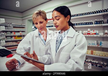 Giovane donna che lavora in farmacia mostrando tablet digitale a. collega senior in piedi dietro il bancone Foto Stock