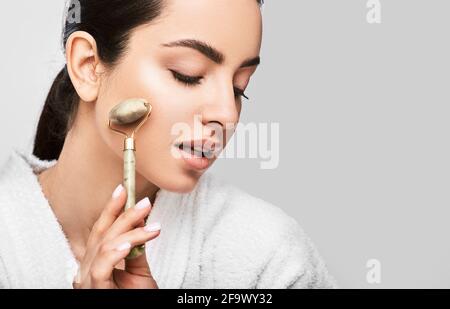 rullo di giada facciale. Una giovane donna massaggia il viso con un rullo di giada verde per una pelle elastica perfetta. Isolato su grigio Foto Stock