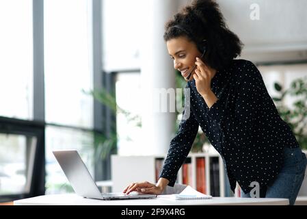 Occupato giovane donna d'affari afro-americana di successo, manager o consulente, in una cuffia, si trova vicino a un desktop in un ufficio, utilizza un computer portatile, digitando un messaggio, o comunicando con un cliente online Foto Stock