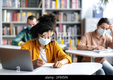 Imparare durante una pandemia. Gli studenti in maschere mediche protettive siedono nella biblioteca universitaria a distanza l'uno dall'altro. Una studentessa afroamericana prende appunti durante la lezione Foto Stock