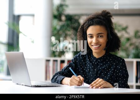 Giovane donna afro-americana di successo, in una cuffia, freelance, dipendente o manager, si siede a un tavolo da lavoro con un laptop e un notebook, prende appunti, guarda la fotocamera, sorride amichevole Foto Stock