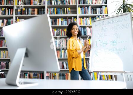 Una piacevole insegnante di afroamericana conduce lezioni online per gli studenti tramite videoconferenza via computer, si trova presso la biblioteca universitaria, mostra informazioni sulla lavagna, guarda il computer, sorride Foto Stock