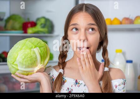 Bella giovane ragazza teen che tiene fresco cavolo verde mentre in piedi vicino frigorifero aperto in cucina a casa Foto Stock